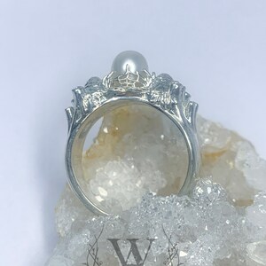 May include: A silver ring with a pearl set in a delicate, intricate design. The ring is on a bed of white crystals.