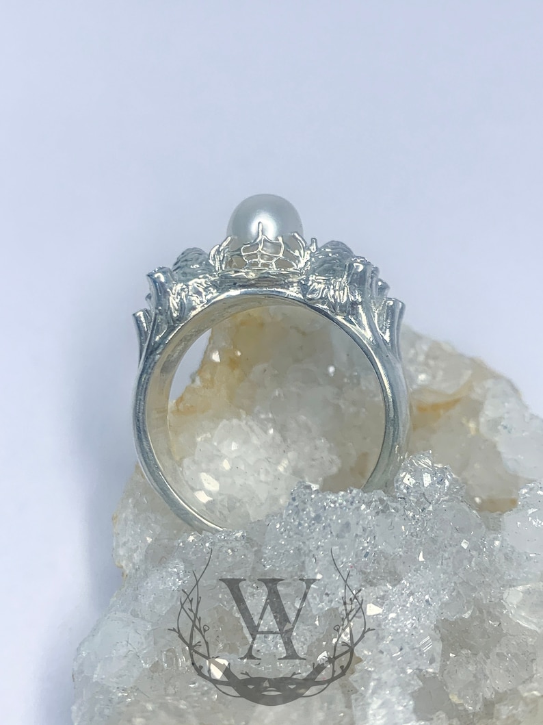 May include: A silver ring with a pearl set in a delicate, intricate design. The ring is on a bed of white crystals.