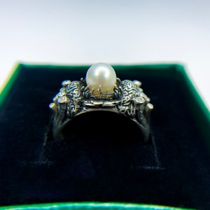 May include: A silver ring with a large pearl set in a detailed design. The ring is in a green velvet box.