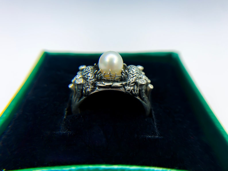 May include: A silver ring with a large pearl set in a detailed design. The ring is in a green velvet box.
