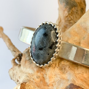May include: A silver bangle bracelet with a black oval gemstone set in a silver bezel with a decorative border. The bracelet is resting on a piece of driftwood.