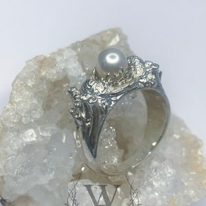 May include: A silver ring with a pearl set in a detailed, textured design. The ring is displayed on a white crystal surface.