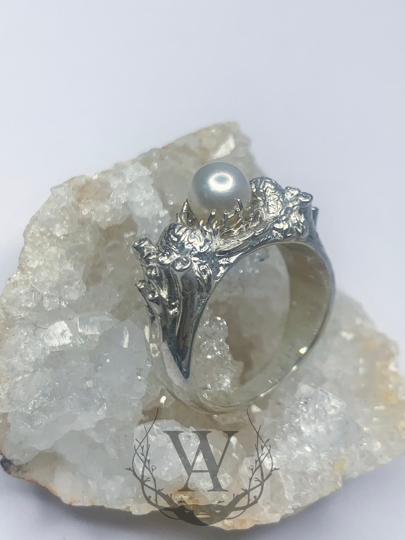 May include: A silver ring with a pearl set in a detailed, textured design. The ring is displayed on a white crystal surface.