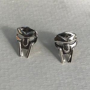 May include: A pair of silver-colored stud earrings. Each earring is shaped like a stylized mask with a rectangular top, eye openings, and a vertical bar extending downward. The earrings are displayed on a light-colored surface.