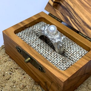 May include: A silver ring with a large pearl and intricate details, displayed in a wooden box with a burlap lining.