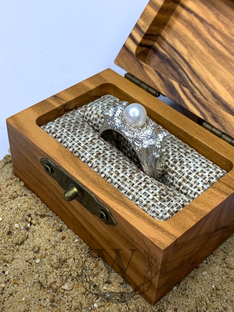 May include: A silver ring with a large pearl and intricate details, displayed in a wooden box with a burlap lining.