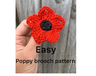 May include: A red crocheted poppy flower with a black button center. The flower is held in a hand and the text "Easy Poppy brooch pattern" is below the flower.