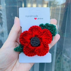 Könnte beinhalten: Eine handgefertigte rote Häkel-Mohnblume mit schwarzem Knopf in der Mitte und grünen Blättern. Die Mohnblume ist an einer weißen Karte befestigt, auf der "Handmade with love" steht.
