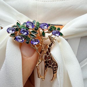 K&ouml;nnte beinhalten: Goldfarbene Brosche mit einem Baumdesign, lila Edelsteinakzenten und einer Giraffe. Die Brosche hat gr&uuml;ne Blattdetails und ist vor einem wei&szlig;en Stoffhintergrund platziert.