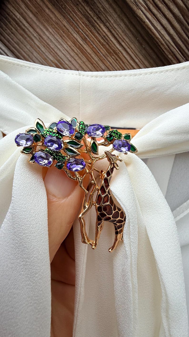K&ouml;nnte beinhalten: Goldfarbene Brosche mit einem Baumdesign, lila Edelsteinakzenten und einer Giraffe. Die Brosche hat gr&uuml;ne Blattdetails und ist vor einem wei&szlig;en Stoffhintergrund platziert.