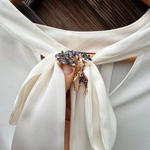 K&ouml;nnte beinhalten: Goldfarbene Brosche mit Giraffen-Design und einem Baum, der mit lila und gr&uuml;nen Edelsteinen verziert ist. Die Brosche ist an einer wei&szlig;en Bluse mit Schleife befestigt. Dekoratives Accessoire.