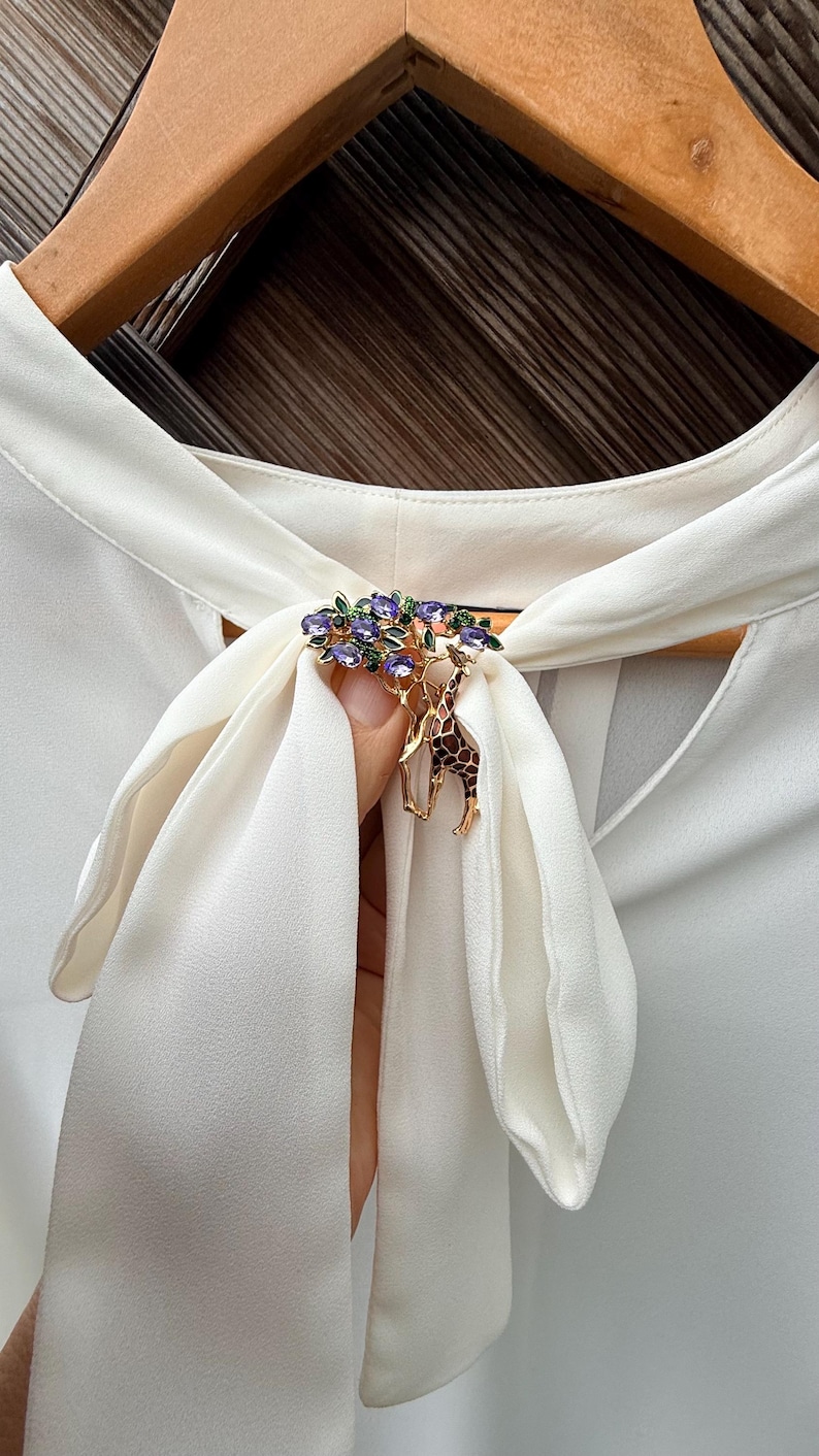 K&ouml;nnte beinhalten: Goldfarbene Brosche mit Giraffen-Design und einem Baum, der mit lila und gr&uuml;nen Edelsteinen verziert ist. Die Brosche ist an einer wei&szlig;en Bluse mit Schleife befestigt. Dekoratives Accessoire.
