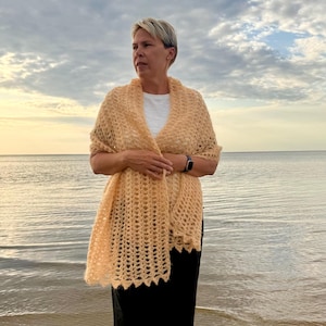 Könnte beinhalten: Eine Frau trägt einen pfirsichfarbenen, offen gewebten Häkelschal. Der Schal ist über den Schultern drapiert und hat einen gewellten Rand. Sie steht an einem Strand mit dem Meer und einem bewölkten Himmel im Hintergrund.