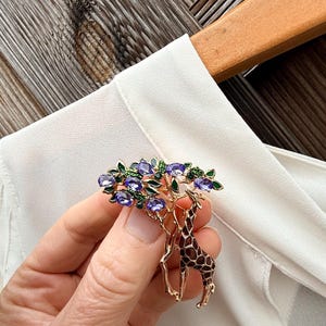 K&ouml;nnte beinhalten: Eine goldfarbene Brosche mit einem Giraffen-Design und einem Baum. Der Baum hat lila Edelsteinbl&auml;tter und gr&uuml;ne Akzente. Die Giraffe hat ein braunes und beiges Muster. Die Brosche wird vor einer wei&szlig;en Bluse gehalten.