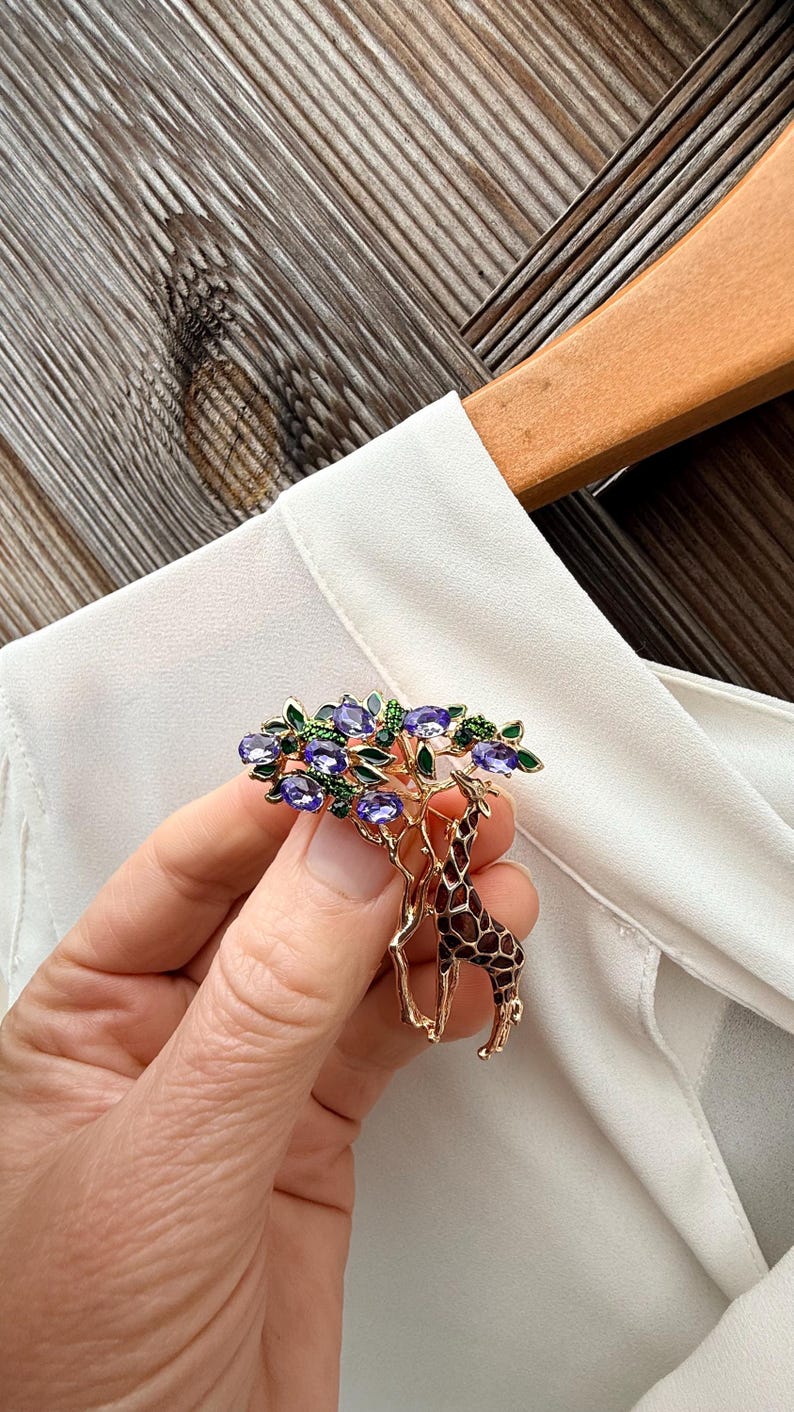K&ouml;nnte beinhalten: Eine goldfarbene Brosche mit einem Giraffen-Design und einem Baum. Der Baum hat lila Edelsteinbl&auml;tter und gr&uuml;ne Akzente. Die Giraffe hat ein braunes und beiges Muster. Die Brosche wird vor einer wei&szlig;en Bluse gehalten.