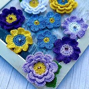 May include: A collection of crocheted flowers in various colors, including purple, blue, yellow, and white. The flowers are arranged in a silver frame on a wooden surface.