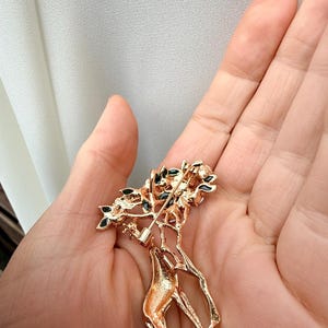 K&ouml;nnte beinhalten: Goldfarbene Brosche in Form einer Giraffe mit einem Baum auf dem R&uuml;cken. Der Baum hat gr&uuml;ne und schwarze Akzente. Die Brosche wird in der Hand einer Person gehalten.