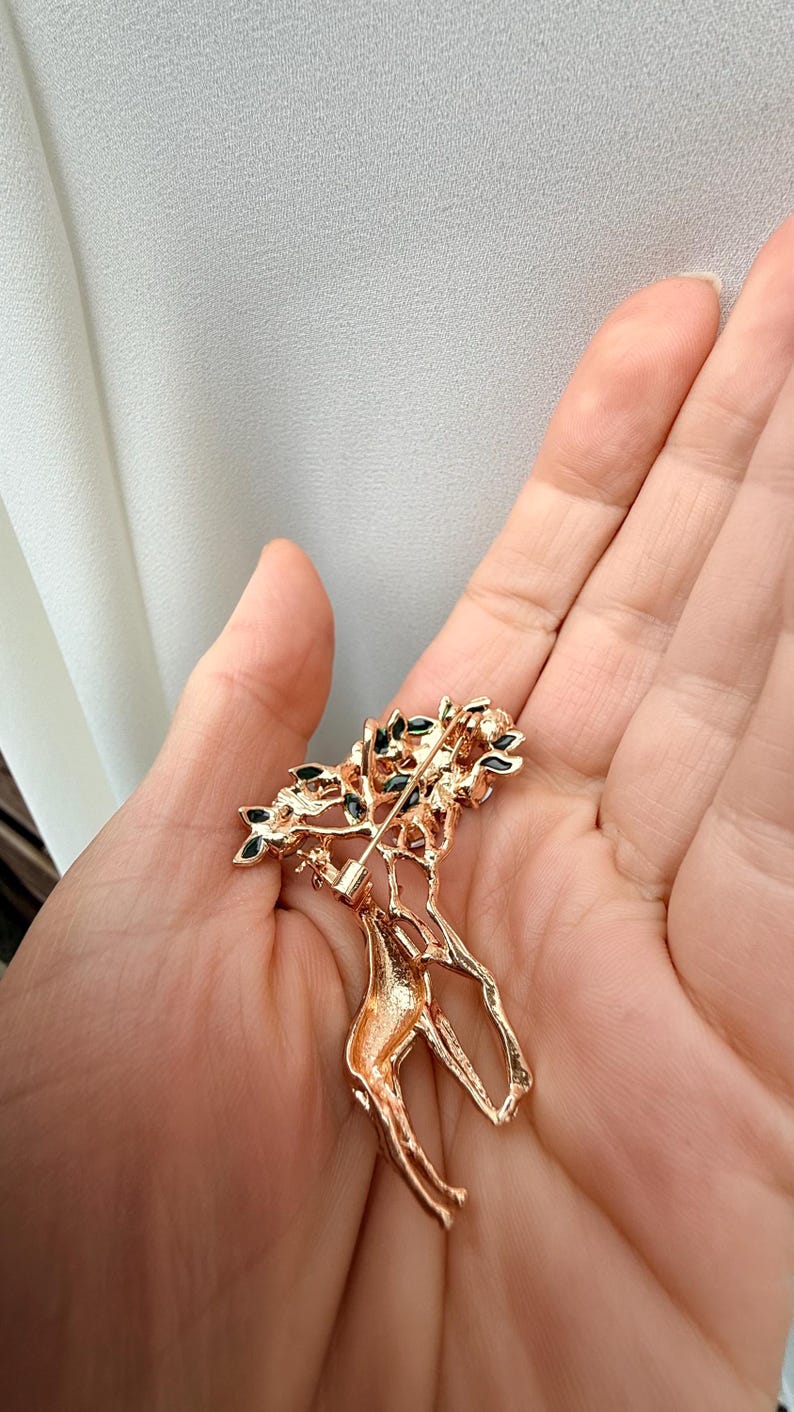 K&ouml;nnte beinhalten: Goldfarbene Brosche in Form einer Giraffe mit einem Baum auf dem R&uuml;cken. Der Baum hat gr&uuml;ne und schwarze Akzente. Die Brosche wird in der Hand einer Person gehalten.