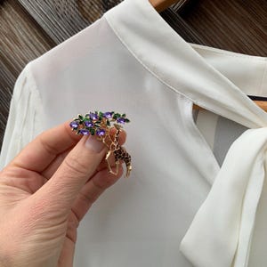 K&ouml;nnte beinhalten: Goldfarbene Brosche mit Giraffen-Design und baumartiger Struktur, verziert mit lila Edelsteinen und gr&uuml;nen Bl&auml;ttern. Die Giraffe hat braune und schwarze Flecken. Die Brosche wird vor einer wei&szlig;en Bluse gehalten.