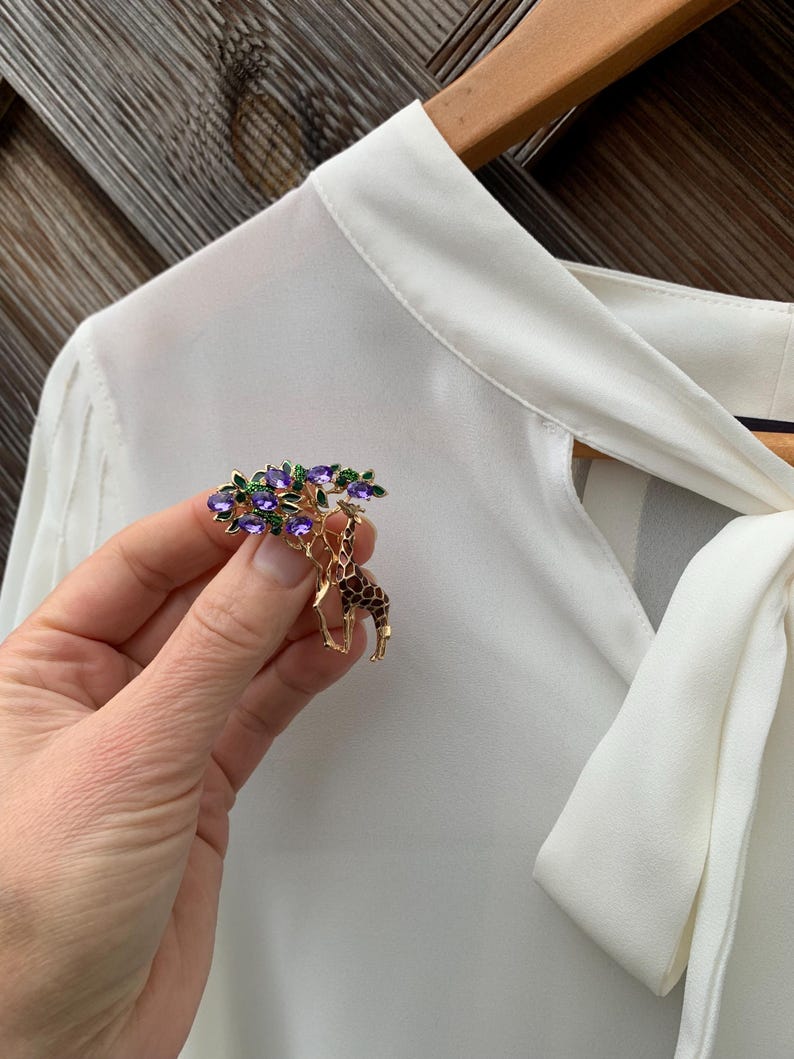 K&ouml;nnte beinhalten: Goldfarbene Brosche mit Giraffen-Design und baumartiger Struktur, verziert mit lila Edelsteinen und gr&uuml;nen Bl&auml;ttern. Die Giraffe hat braune und schwarze Flecken. Die Brosche wird vor einer wei&szlig;en Bluse gehalten.