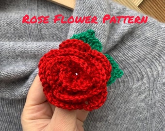 May include: A crocheted red rose with green leaves. The rose is attached to a gray knitted sweater. The text "ROSE FLOWER PATTERN" is written above the rose.