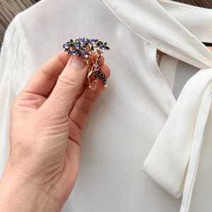 K&ouml;nnte beinhalten: Goldfarbene Brosche mit Giraffen-Design unter einem Baum. Der Baum hat lila und gr&uuml;ne Akzente. Die Giraffe hat ein braunes und schwarzes Muster. Die Brosche wird vor einer wei&szlig;en Bluse gehalten.