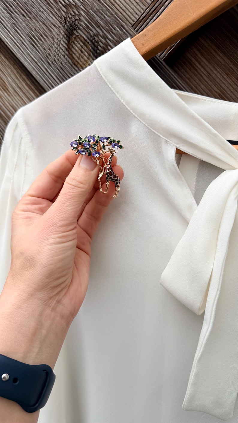 K&ouml;nnte beinhalten: Goldfarbene Brosche mit Giraffen-Design unter einem Baum. Der Baum hat lila und gr&uuml;ne Akzente. Die Giraffe hat ein braunes und schwarzes Muster. Die Brosche wird vor einer wei&szlig;en Bluse gehalten.