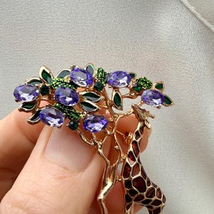 K&ouml;nnte beinhalten: Goldfarbene Brosche mit einem Giraffen- und Baum-Design. Der Baum hat lila, ovale Edelsteine und gr&uuml;ne Akzente. Die Giraffe hat ein braunes und rotes Muster. Die Brosche wird von einer Hand gehalten.