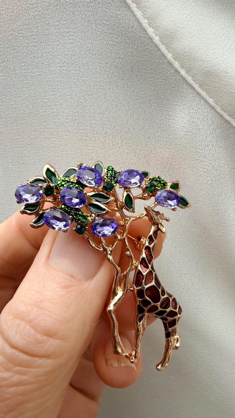 K&ouml;nnte beinhalten: Goldfarbene Brosche mit einem Giraffen- und Baum-Design. Der Baum hat lila, ovale Edelsteine und gr&uuml;ne Akzente. Die Giraffe hat ein braunes und rotes Muster. Die Brosche wird von einer Hand gehalten.