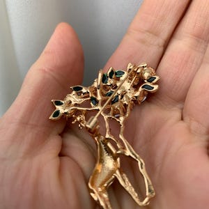 K&ouml;nnte beinhalten: Goldfarbene Brosche in Form eines Hirsches mit floralem Design. Der Hirsch hat einen detaillierten, strukturierten K&ouml;rper und ist mit kleinen, dunkelgr&uuml;nen Akzenten auf den floralen Elementen verziert. Die Brosche wird in einer Hand gehalten.