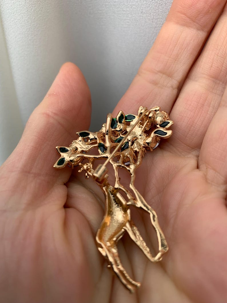 K&ouml;nnte beinhalten: Goldfarbene Brosche in Form eines Hirsches mit floralem Design. Der Hirsch hat einen detaillierten, strukturierten K&ouml;rper und ist mit kleinen, dunkelgr&uuml;nen Akzenten auf den floralen Elementen verziert. Die Brosche wird in einer Hand gehalten.