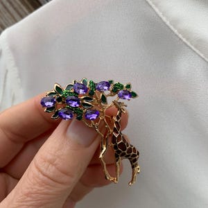 K&ouml;nnte beinhalten: Goldfarbene Brosche mit Giraffen- und Baumdesign. Der Baum hat violette und gr&uuml;ne Edelsteinakzente. Die Giraffe hat ein braunes und schwarzes Muster. Die Brosche wird vor einem wei&szlig;en Hintergrund gehalten.