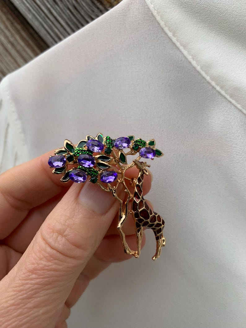 K&ouml;nnte beinhalten: Goldfarbene Brosche mit Giraffen- und Baumdesign. Der Baum hat violette und gr&uuml;ne Edelsteinakzente. Die Giraffe hat ein braunes und schwarzes Muster. Die Brosche wird vor einem wei&szlig;en Hintergrund gehalten.