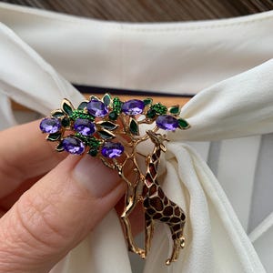 K&ouml;nnte beinhalten: Goldfarbene Brosche mit einer Giraffe und einem Baum, verziert mit lila und gr&uuml;nen Edelsteinen. Die Giraffe hat ein braunes und beiges Muster. Die Brosche befindet sich vor einem wei&szlig;en Stoffhintergrund.