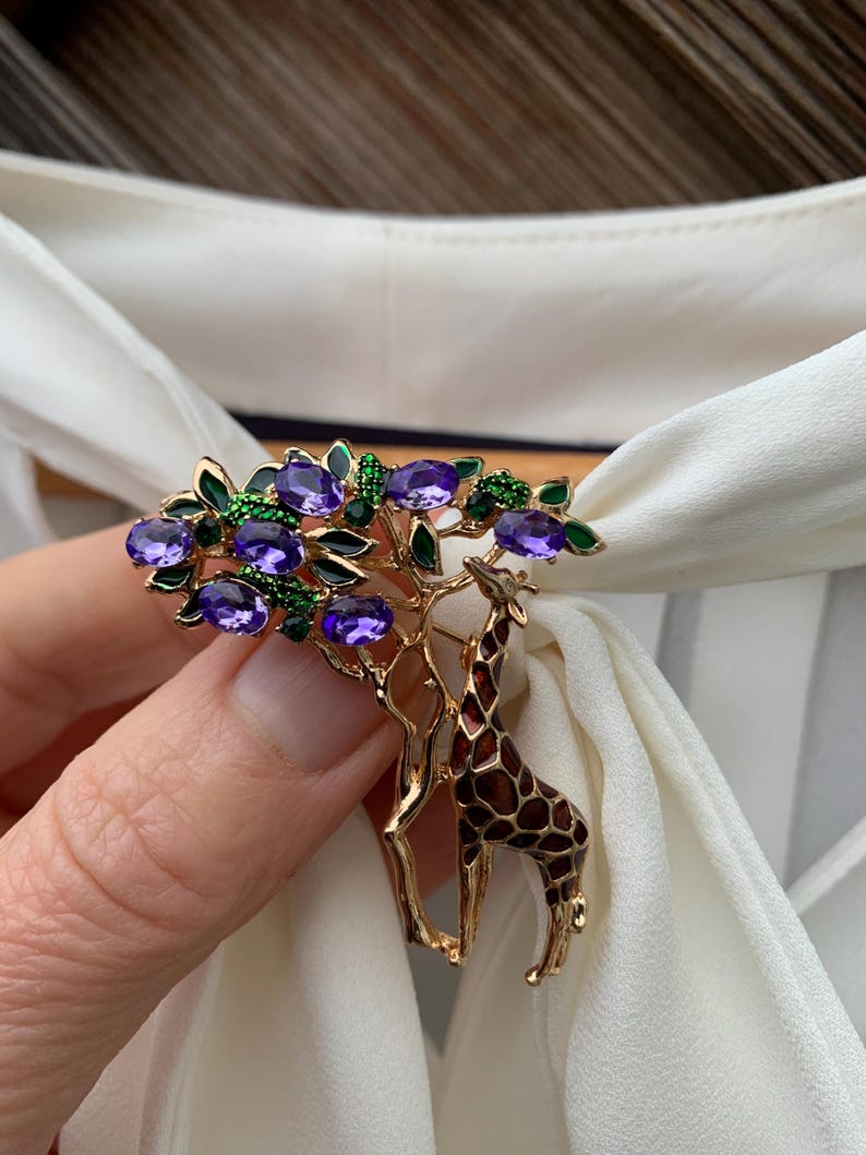 K&ouml;nnte beinhalten: Goldfarbene Brosche mit einer Giraffe und einem Baum, verziert mit lila und gr&uuml;nen Edelsteinen. Die Giraffe hat ein braunes und beiges Muster. Die Brosche befindet sich vor einem wei&szlig;en Stoffhintergrund.