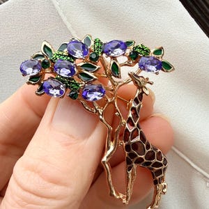 K&ouml;nnte beinhalten: Goldfarbene Brosche mit einer Giraffe unter einem Baum mit lila Edelsteinbl&auml;ttern und gr&uuml;nen Akzenten. Die Giraffe hat ein braunes und beiges Muster. Die Brosche wird vor einem wei&szlig;en Hintergrund gehalten.