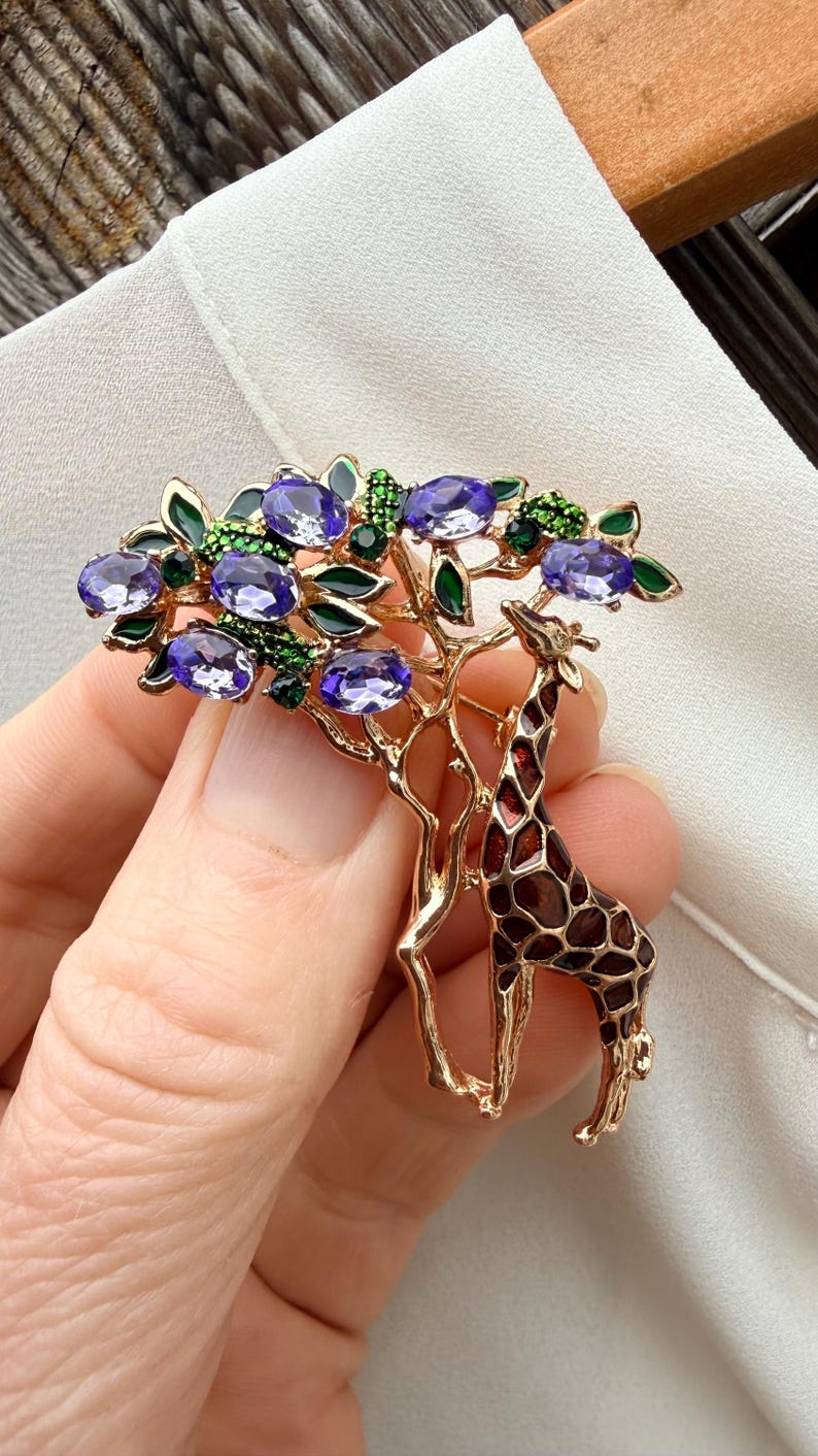 K&ouml;nnte beinhalten: Goldfarbene Brosche mit einer Giraffe unter einem Baum mit lila Edelsteinbl&auml;ttern und gr&uuml;nen Akzenten. Die Giraffe hat ein braunes und beiges Muster. Die Brosche wird vor einem wei&szlig;en Hintergrund gehalten.