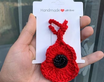 May include: A handmade red crochet poppy flower with a black center, attached to a white card. The card reads "Handmade with love". The poppy is a symbol of remembrance.