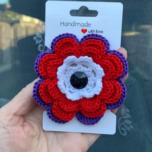 Könnte beinhalten: Handgefertigte Häkelblumenbrosche mit roten, weißen und lila Blütenblättern. Die Mitte hat einen schwarzen Edelstein. Auf der Karte steht "Handmade with love."