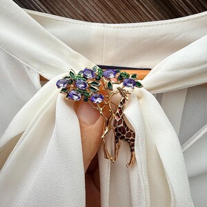 K&ouml;nnte beinhalten: Goldfarbene Brosche mit Giraffen-Design und einem Baum mit lila Edelsteinen und gr&uuml;nen Bl&auml;ttern. Die Giraffe hat ein braunes und beiges Muster. Die Brosche ist vor einem wei&szlig;en Stoff platziert.