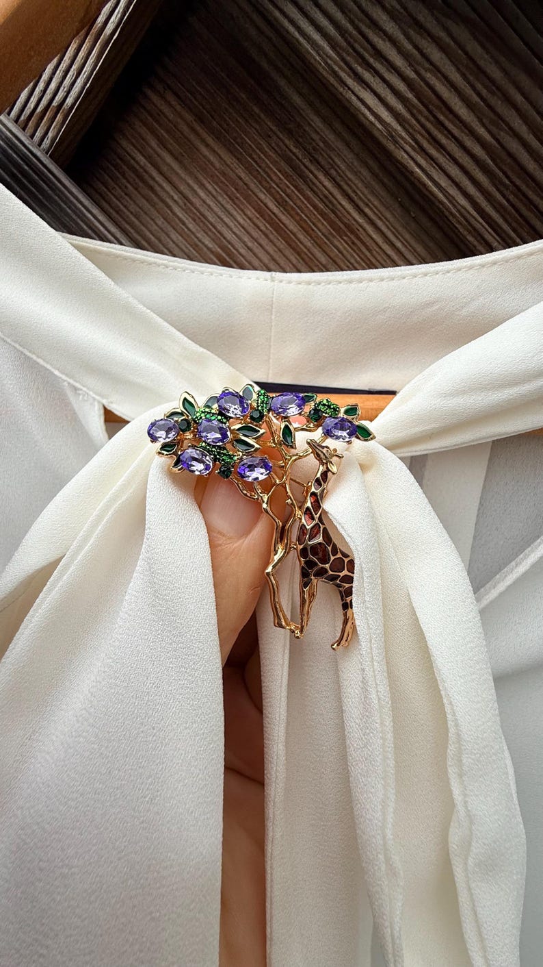 K&ouml;nnte beinhalten: Goldfarbene Brosche mit Giraffen-Design und einem Baum mit lila Edelsteinen und gr&uuml;nen Bl&auml;ttern. Die Giraffe hat ein braunes und beiges Muster. Die Brosche ist vor einem wei&szlig;en Stoff platziert.
