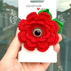 Könnte beinhalten: Eine gehäkelte rote Mohnblumen-Brosche mit grünen Blättern, die in der Mitte eine schwarze facettierte Perle umgeben von kleinen weißen Saatperlen aufweist. Die Blume ist auf einer weißen rechteckigen Karte mit dem Text Handmade with love befestigt.