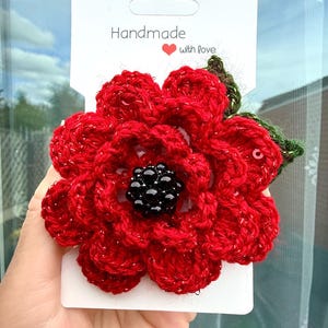 Könnte beinhalten: Eine gehäkelte rote Blumenbrosche mit schwarzer Perlenmitte und einem kleinen grünen Blatt, befestigt auf einer weißen Präsentationskarte mit dem Text Handmade with love und einem roten Herzsymbol. Das rote Garn enthält feine Metallicfasern.