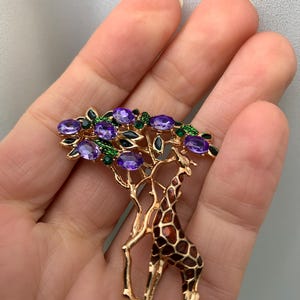 K&ouml;nnte beinhalten: Goldfarbene Brosche mit einer Giraffe unter einem Baum mit lila Edelsteinbl&auml;ttern. Die Giraffe hat ein braun-orangefarbenes Fellmuster. Der Baum hat gr&uuml;ne und schwarze Akzente. Ein einzigartiges und detailliertes Schmuckst&uuml;ck.