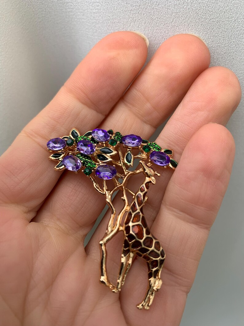 K&ouml;nnte beinhalten: Goldfarbene Brosche mit einer Giraffe unter einem Baum mit lila Edelsteinbl&auml;ttern. Die Giraffe hat ein braun-orangefarbenes Fellmuster. Der Baum hat gr&uuml;ne und schwarze Akzente. Ein einzigartiges und detailliertes Schmuckst&uuml;ck.
