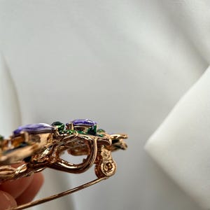 K&ouml;nnte beinhalten: Goldfarbene Brosche mit floralem Design. Die Brosche hat lila und gr&uuml;ne Edelsteine mit aufwendigen Details. Die Nadel ist vor einem wei&szlig;en Hintergrund platziert.