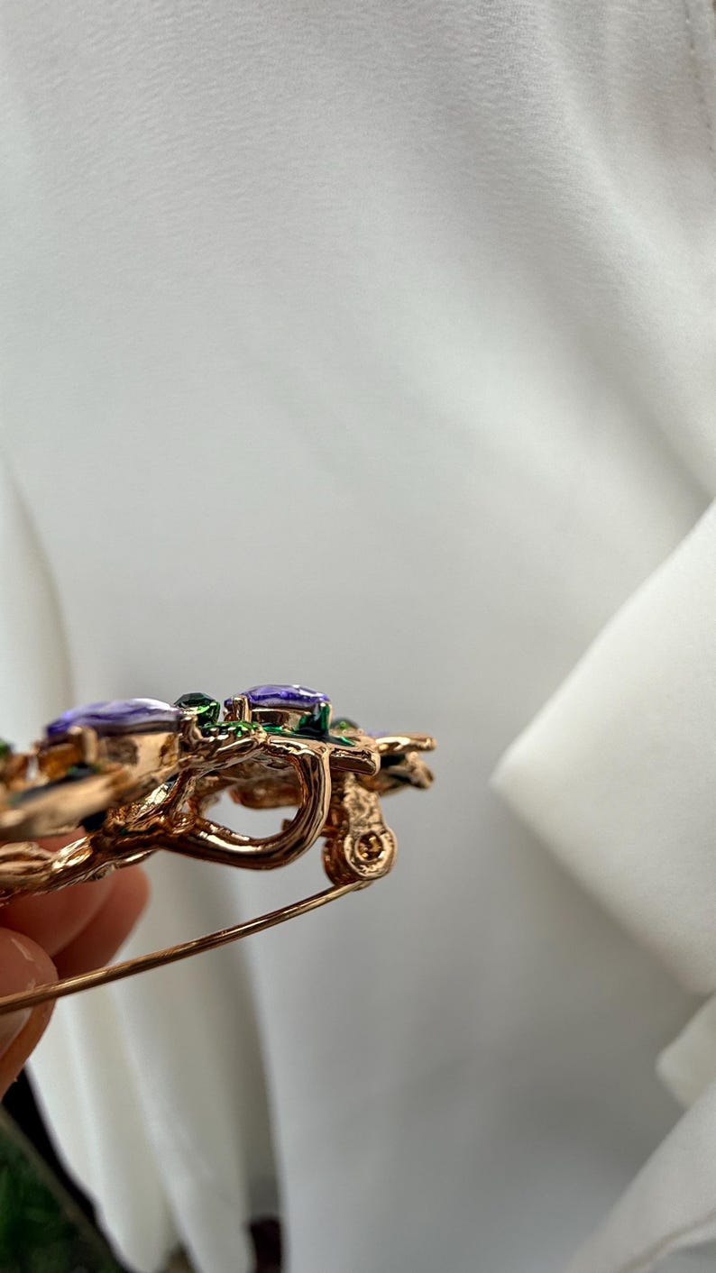 K&ouml;nnte beinhalten: Goldfarbene Brosche mit floralem Design. Die Brosche hat lila und gr&uuml;ne Edelsteine mit aufwendigen Details. Die Nadel ist vor einem wei&szlig;en Hintergrund platziert.