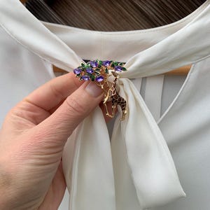 K&ouml;nnte beinhalten: Goldfarbene Brosche mit einem Baumdesign, verziert mit lila und gr&uuml;nen Edelsteinen und einem Giraffenanh&auml;nger. Die Brosche ist an einer wei&szlig;en Bluse befestigt. Ein einzigartiges Accessoire.