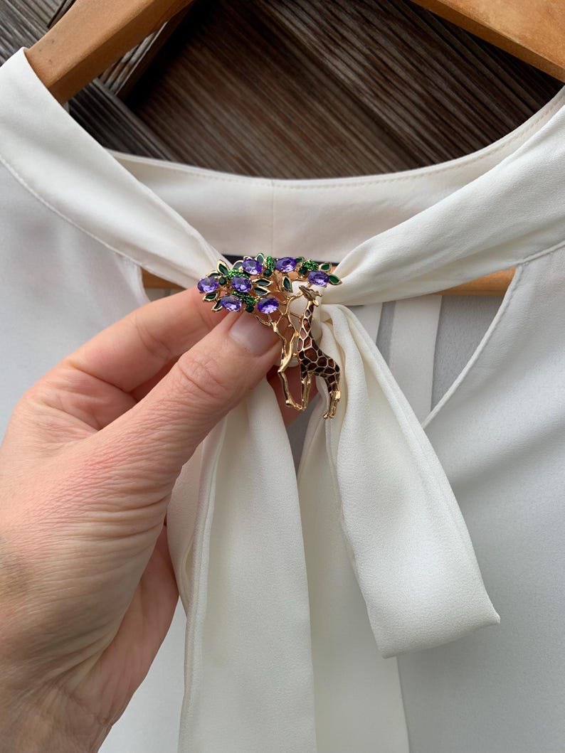 K&ouml;nnte beinhalten: Goldfarbene Brosche mit einem Baumdesign, verziert mit lila und gr&uuml;nen Edelsteinen und einem Giraffenanh&auml;nger. Die Brosche ist an einer wei&szlig;en Bluse befestigt. Ein einzigartiges Accessoire.