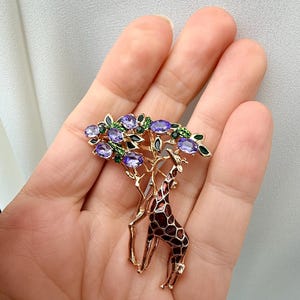 K&ouml;nnte beinhalten: Goldfarbene Brosche mit einer Giraffe unter einem Baum. Der Baum hat violette und gr&uuml;ne Edelsteinakzente. Die Giraffe hat ein braunes und schwarzes Muster. Die Brosche ist ein elegantes Accessoire.
